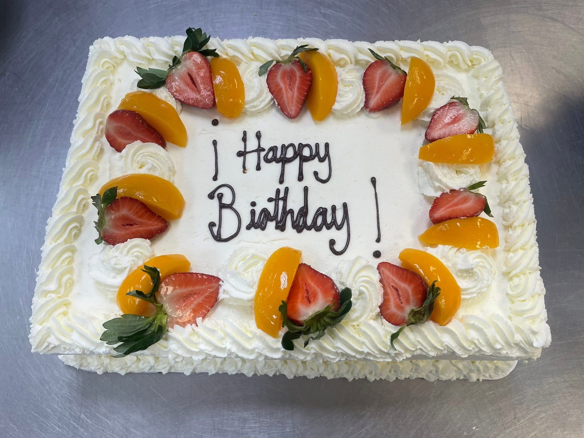 A whipped cream sheet cake piped with Happy Birthday in chocolate, bordered with fresh strawberries and peach slices.