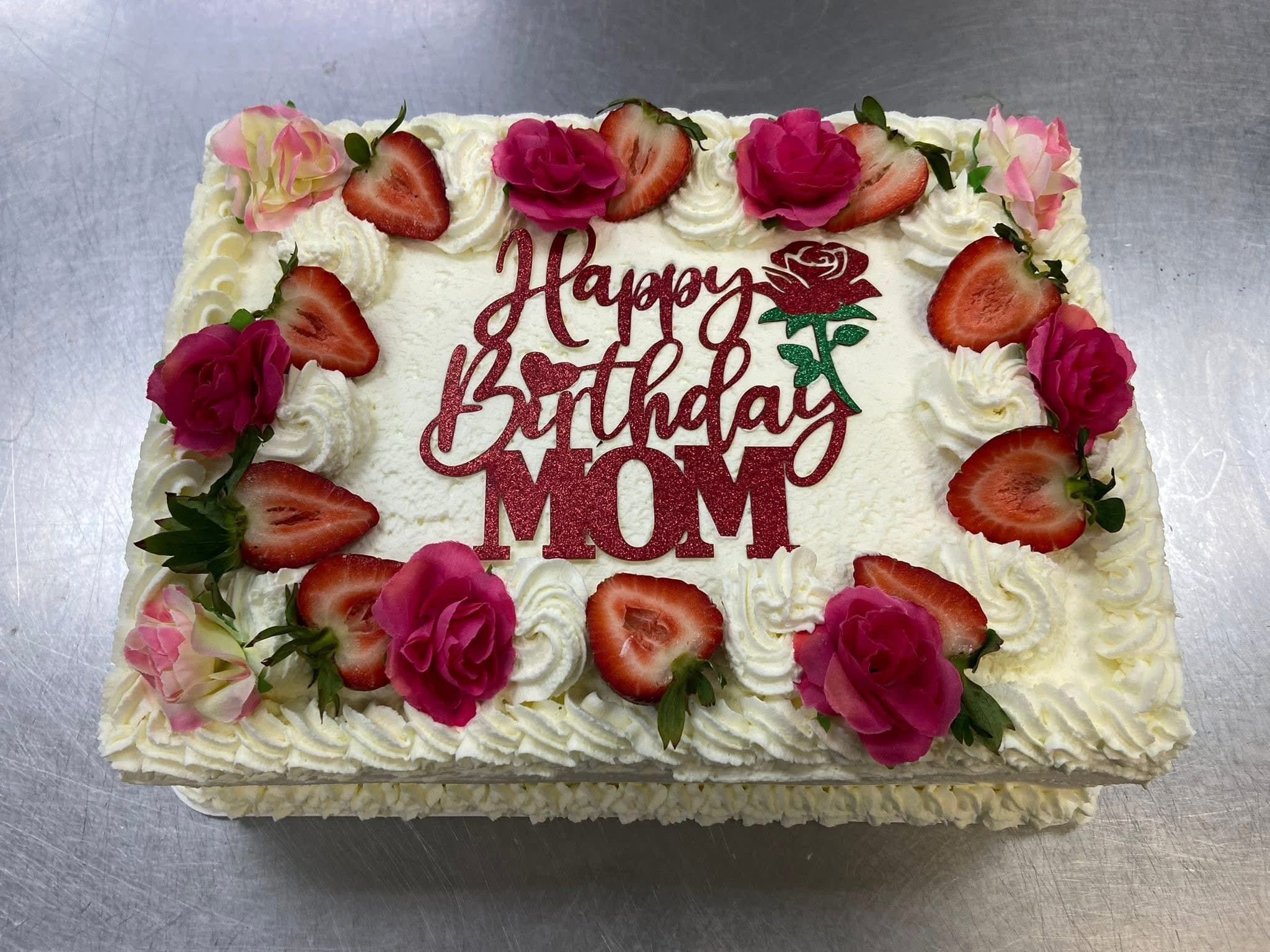 A whipped cream sheet cake with a red Happy Birthday Mom topper, bordered with fresh strawberries and pink roses.