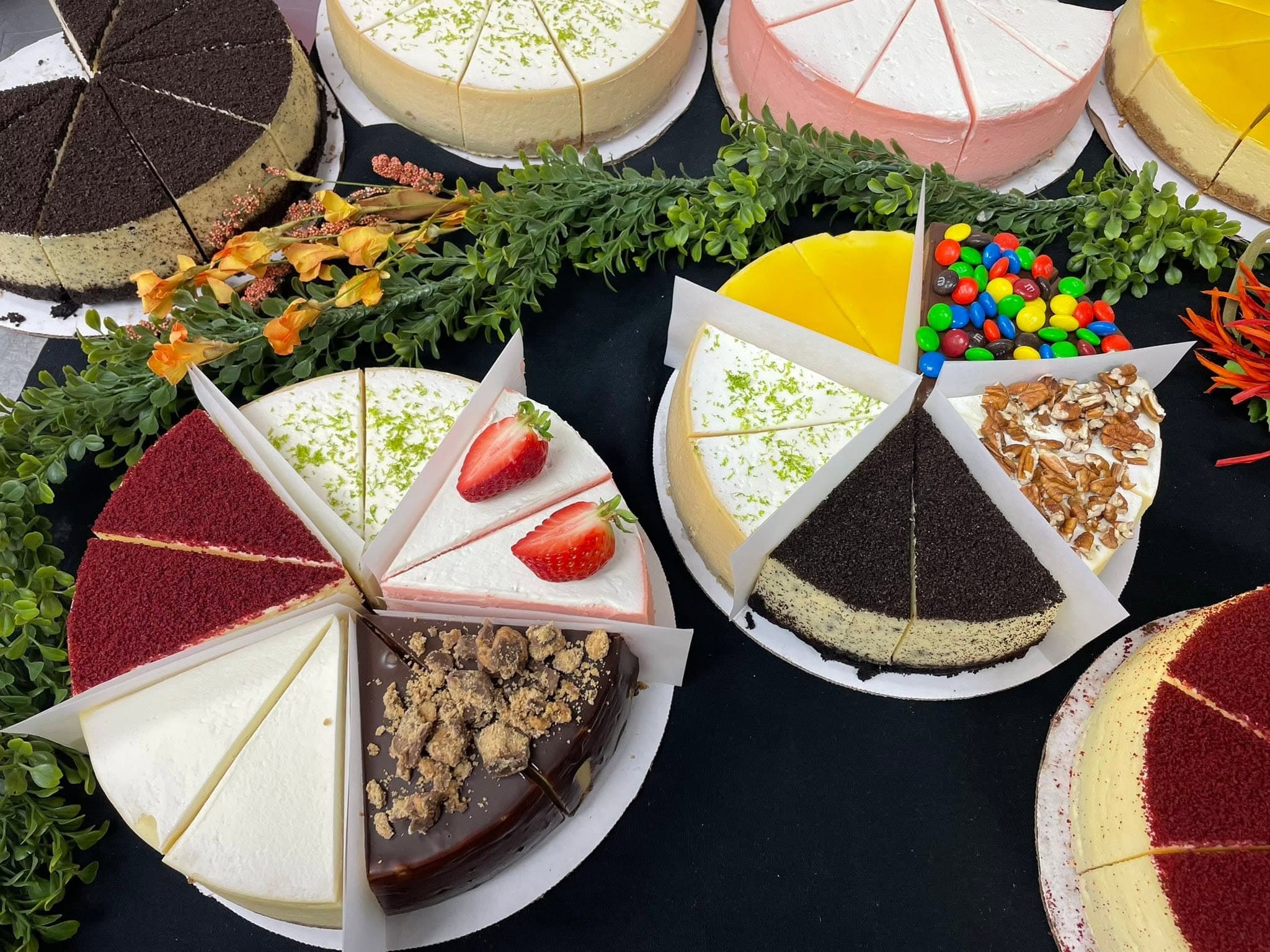 An overhead spread of Yeyo's Bakery cheesecakes — whole cakes cut into mix-and-match slices in flavors including Oreo, key lime, red velvet, strawberry, mango, M&M's, and turtle.
