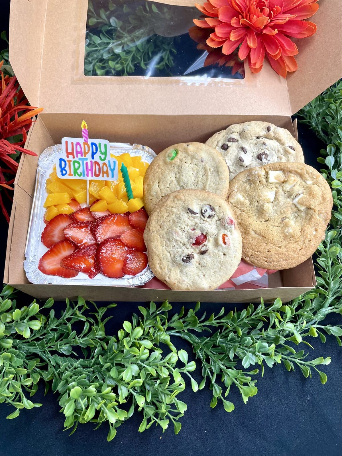 A windowed bakery gift box holding cookies, sliced strawberries and mango, and a lit Happy Birthday candle.