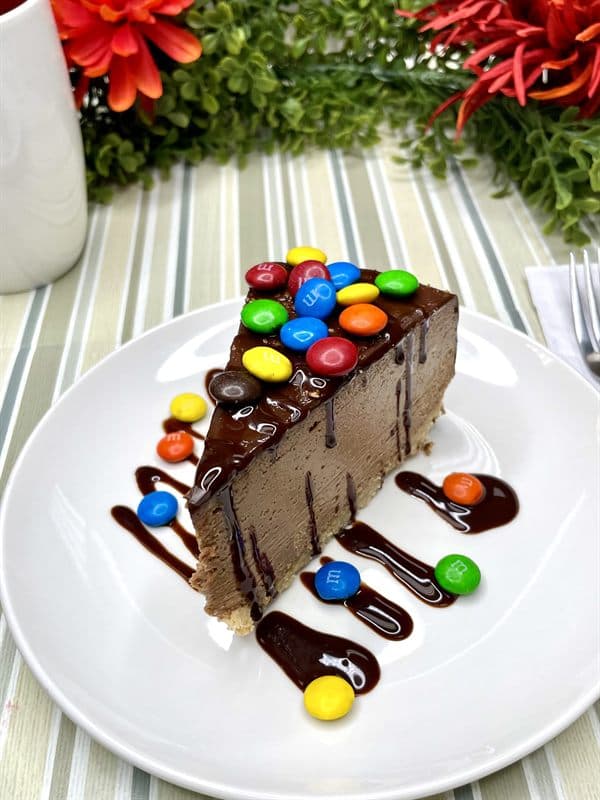 A slice of chocolate cheesecake piled with M&M's and finished with chocolate drizzle.