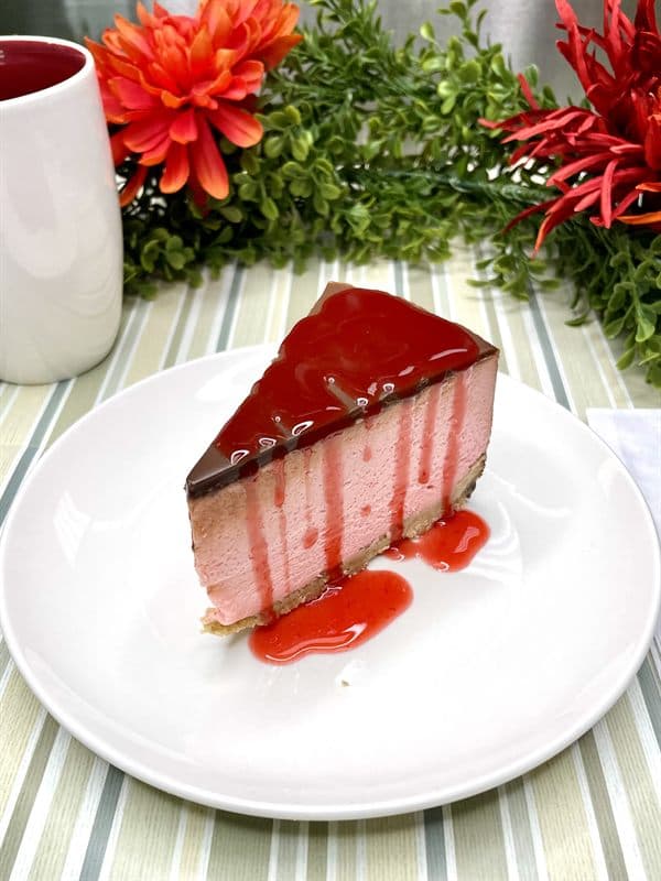 A slice of chocolate covered strawberry cheesecake — pink filling beneath dark chocolate ganache and strawberry sauce.