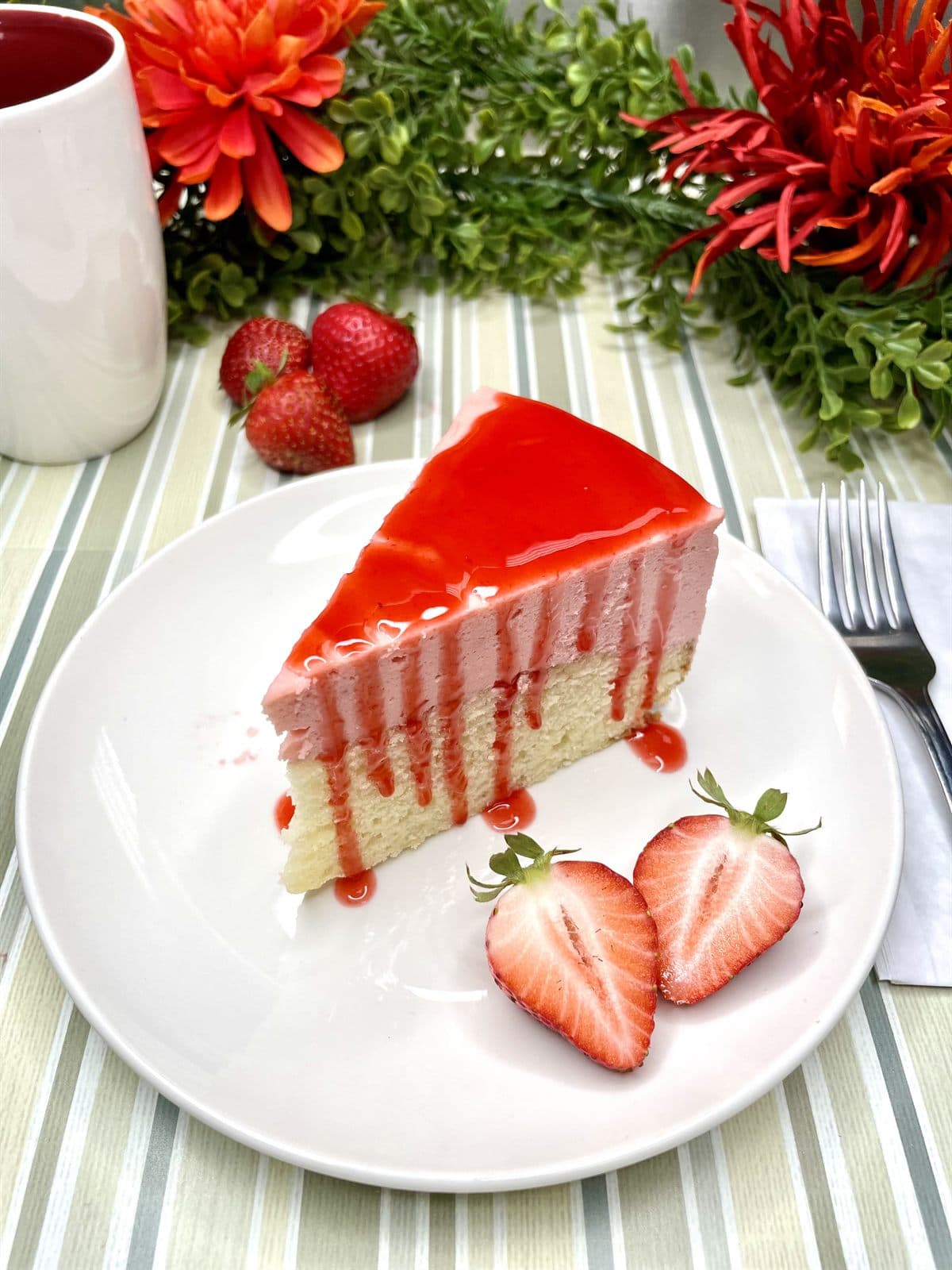 A slice of strawberry cheesecake with a glossy red glaze and fresh strawberries, on a table set with a mug at the bakery.