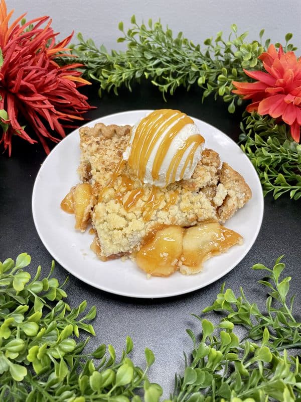 A slice of crumb-topped apple pie with caramel sauce and a scoop of vanilla ice cream.
